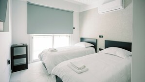 A hotel room with two beds, a small table, a window with a blind, and an air conditioning unit. - Family-Friendly 2BR Apt at Mackenzie Area, Pool available, 10 Min Walk to Beach! (Larnaka)