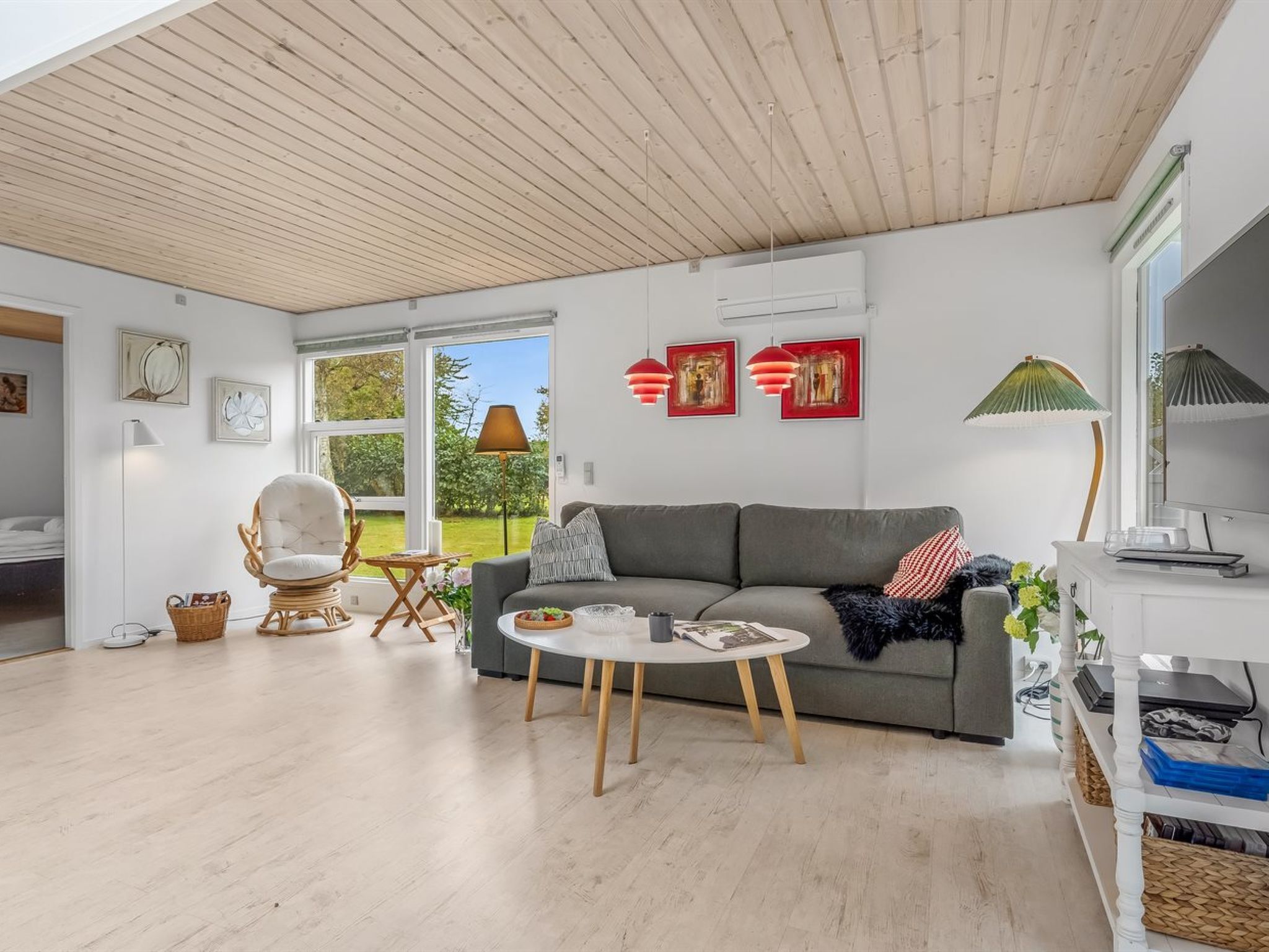 A modern living room with a grey sofa, a round coffee table, and a wicker chair. There are two red pendant lights, a floor lamp, and artwork on the walls.