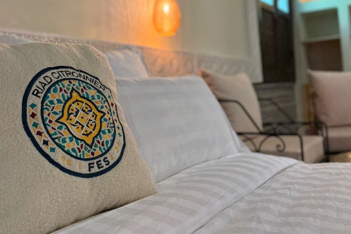 Una cama con un almohadón que presenta un emblema circular, una lámpara colgante y vistas de una habitación con una silla y una mesa.