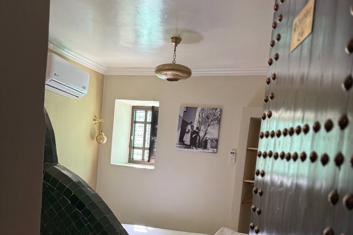 Una habitación con cama, ventana, aire acondicionado de pared y una puerta con diseño metálico decorativo.