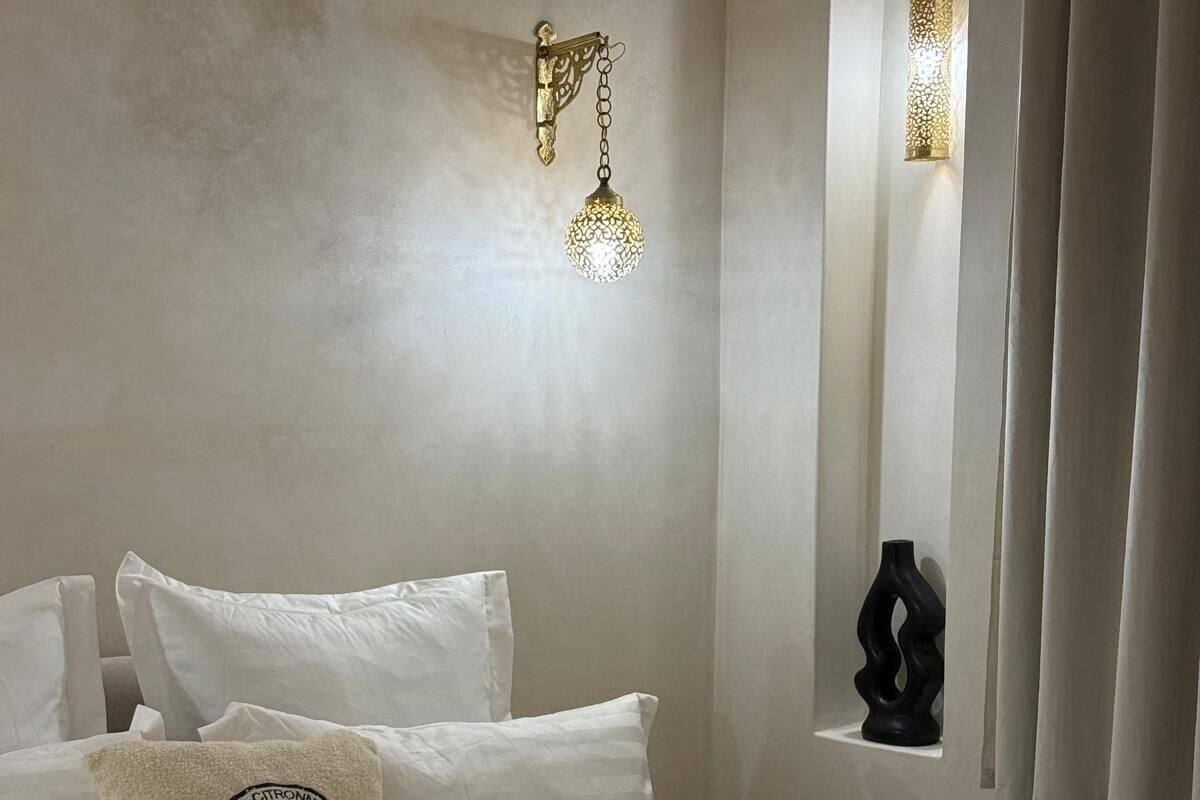 Una cama bien tendida con sábanas blancas y almohadas decorativas, una lámpara de pared y un pequeño ventanal con cortinas.