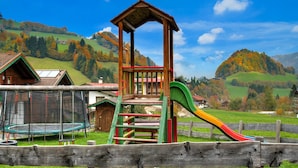 A playground with a slide and swings, surrounded by a wooden fence, set against a backdrop of green hills and a clear sky. - Feriendorf Wildschonau -cozy Escape (Wildschönau)