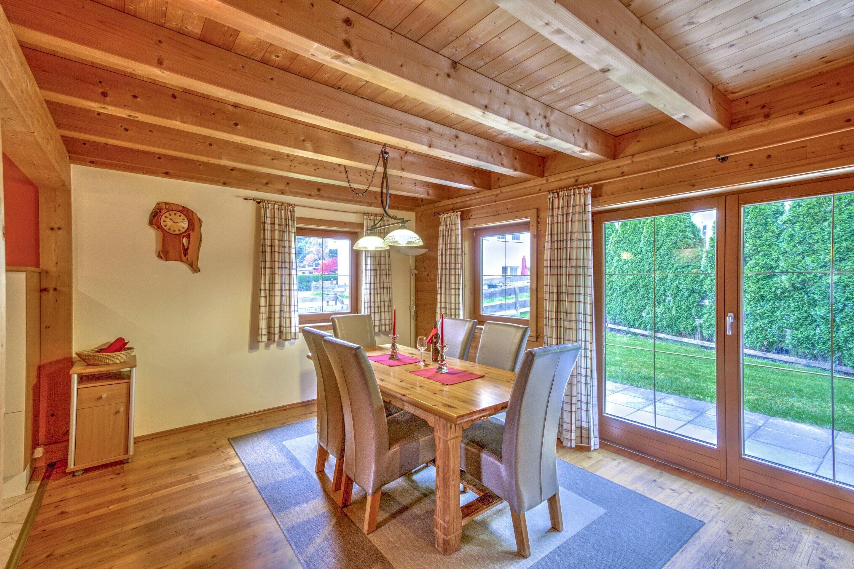 A wooden dining room with a large table, chairs, and a view of a green landscape through glass doors.