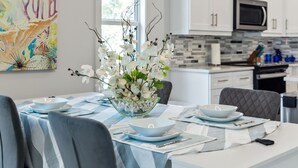 Un espace de salle à manger moderne avec une table dressée pour quatre personnes, un arrangement floral décoratif composé d’une orchidée blanche, et une cuisine en arrière-plan.
