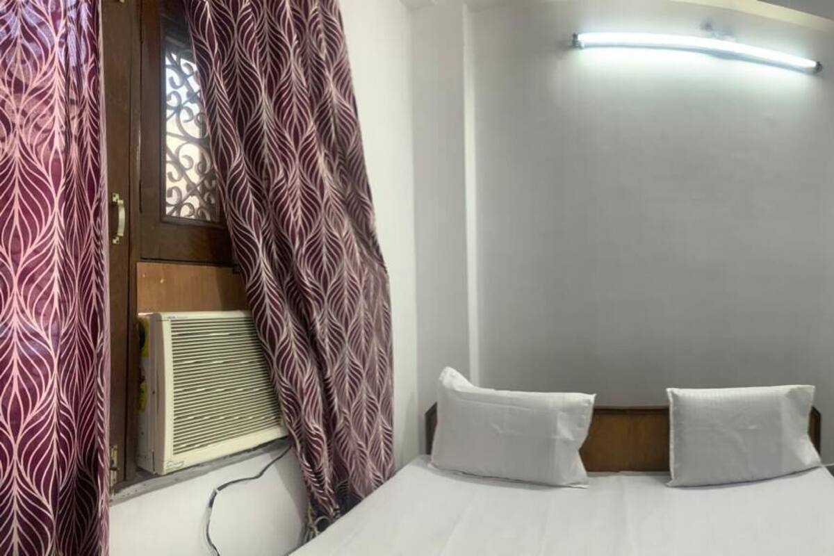 A hotel room with a bed, two pillows, a window with curtains, a wall-mounted TV, and a ceiling light.