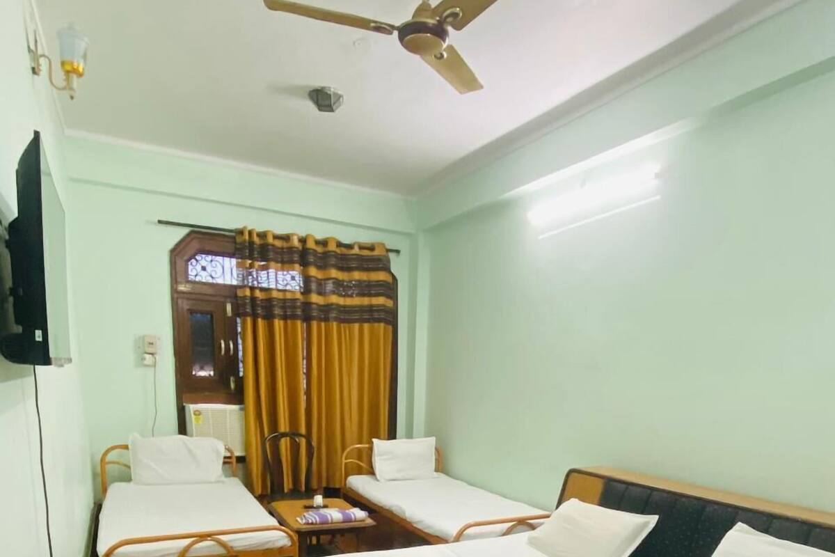 A room with two single beds, a ceiling fan, a window with curtains, and a wall-mounted TV.