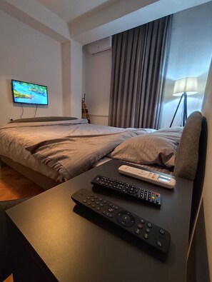 A hotel room with a bed, a TV mounted on the wall, a lamp, and two remote controls on a table.