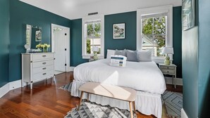 A bedroom with a large bed, a dresser, a bench, and a window with a view of greenery. - The Queens Palace | 8 Bedrooms in Charleston! (Charleston)