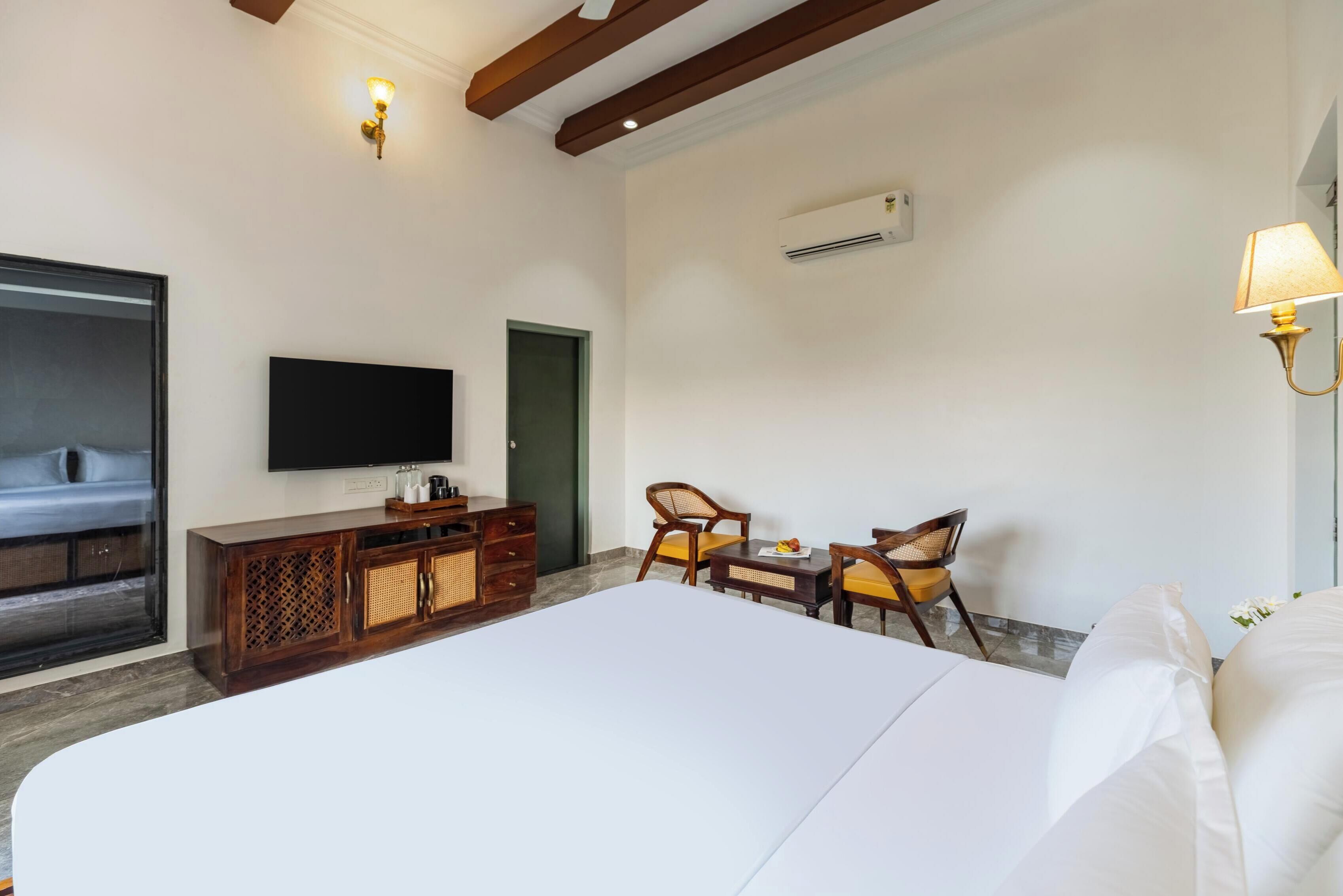 A hotel room with a large bed, a TV mounted on the wall, a wooden cabinet, two chairs, and an air conditioning unit.