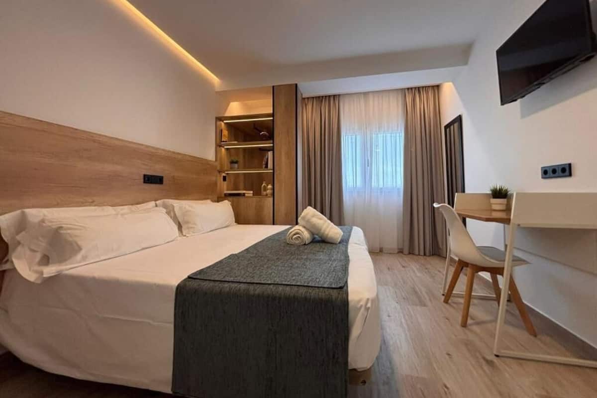 A modern hotel room with a large bed, a desk, a chair, a TV, and a window with curtains.