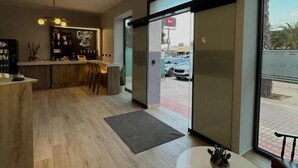 Un hall d’hôtel moderne avec un plancher en bois, une table ronde agrémentée d’une plante en pot, un coin bar avec un néon, et de grandes fenêtres donnant sur la rue.