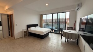 A bedroom with a bed, a desk, and a chair. There is a sliding glass door leading to a balcony with a view of a fence and trees.
