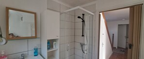 Une salle de bain avec un lavabo blanc, un miroir et une douche. On y trouve des articles de toilette sur le lavabo et sur des étagères.