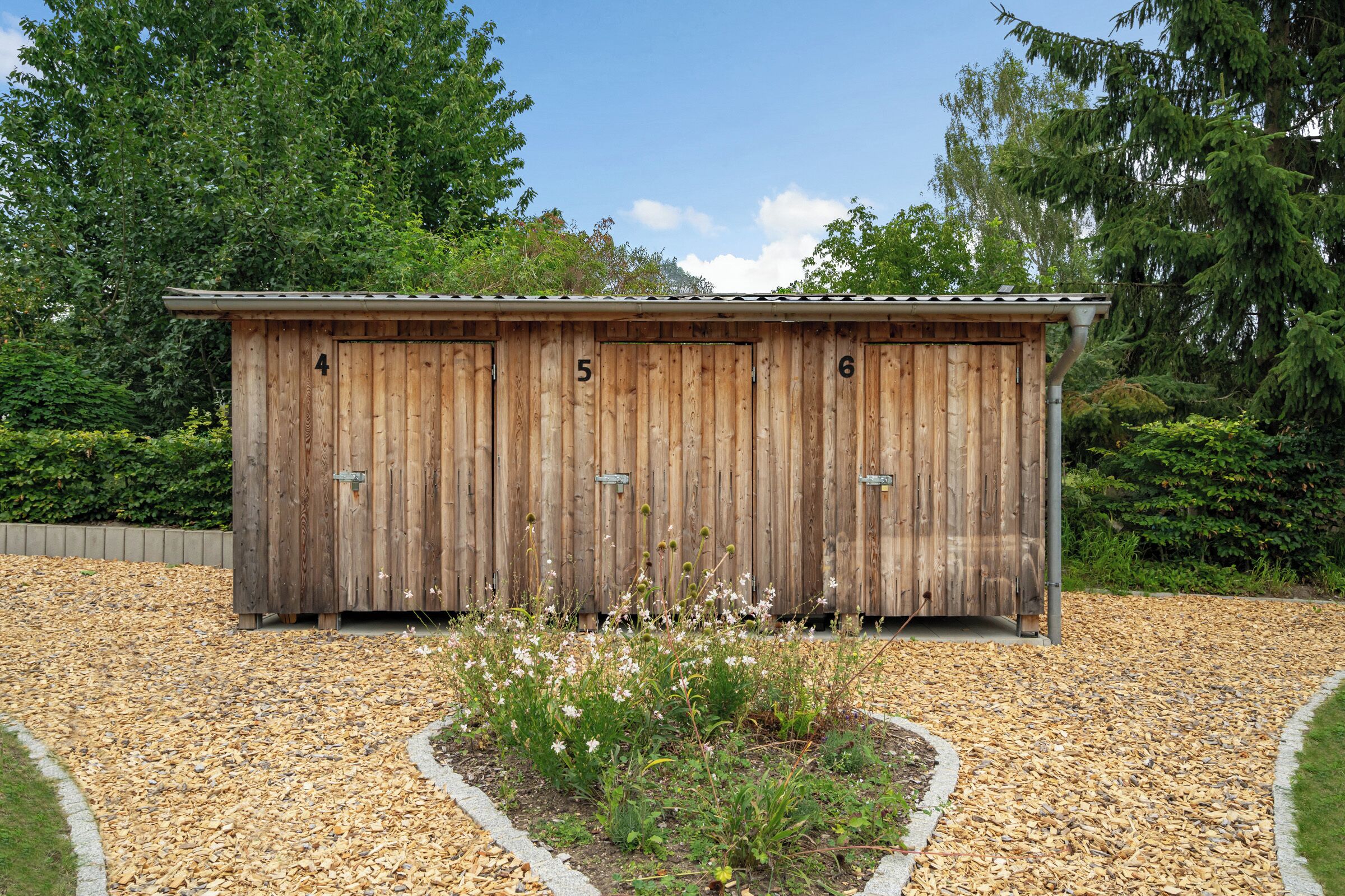 A wooden shed with numbers 4, 5, and 6 on its doors, surrounded by gravel and plants.