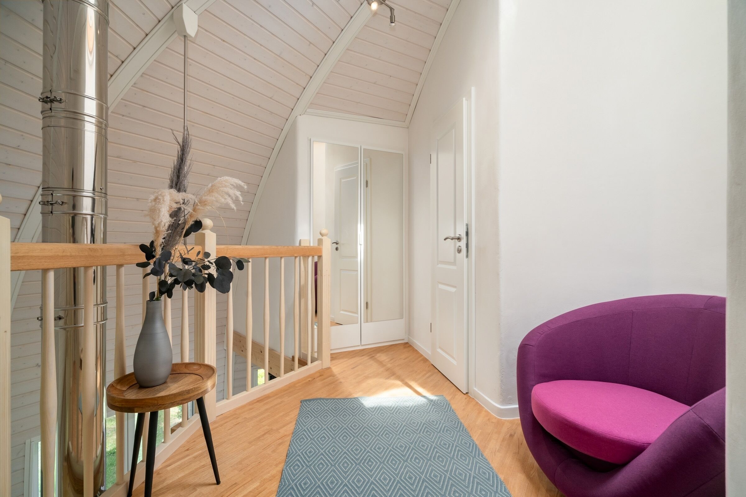 Een moderne loft slaapkamer met een houten plafond, een paarse stoel, een blauw kamerkleed met patroon en een vaas met gedroogde bloemen.