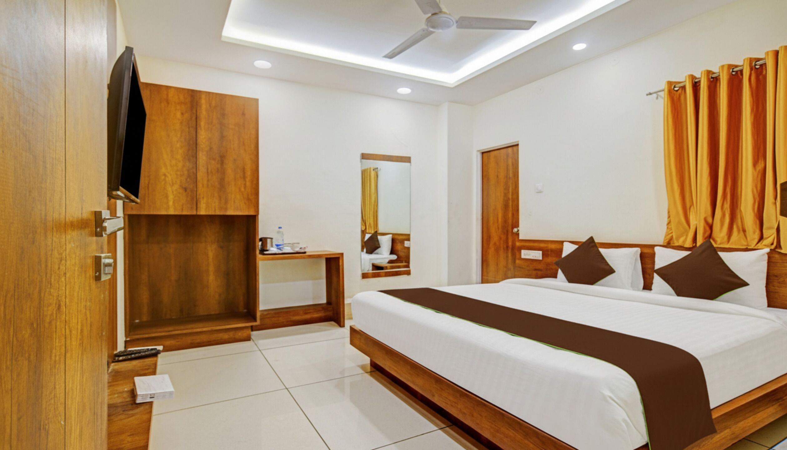 A hotel room with a large bed, wooden wardrobe, a small table with a bottle, a mirror, and a ceiling fan.