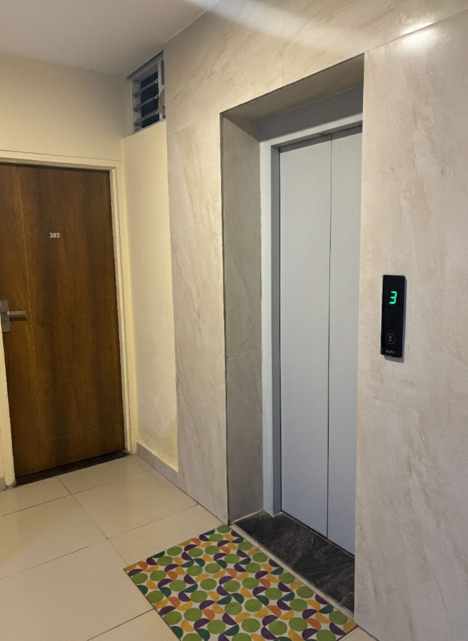 Een lift met een digitaal display waarop het cijfer 3 staat, een houten deur met het nummer 303, en een kleurrijke, patone mat bij de ingang.
