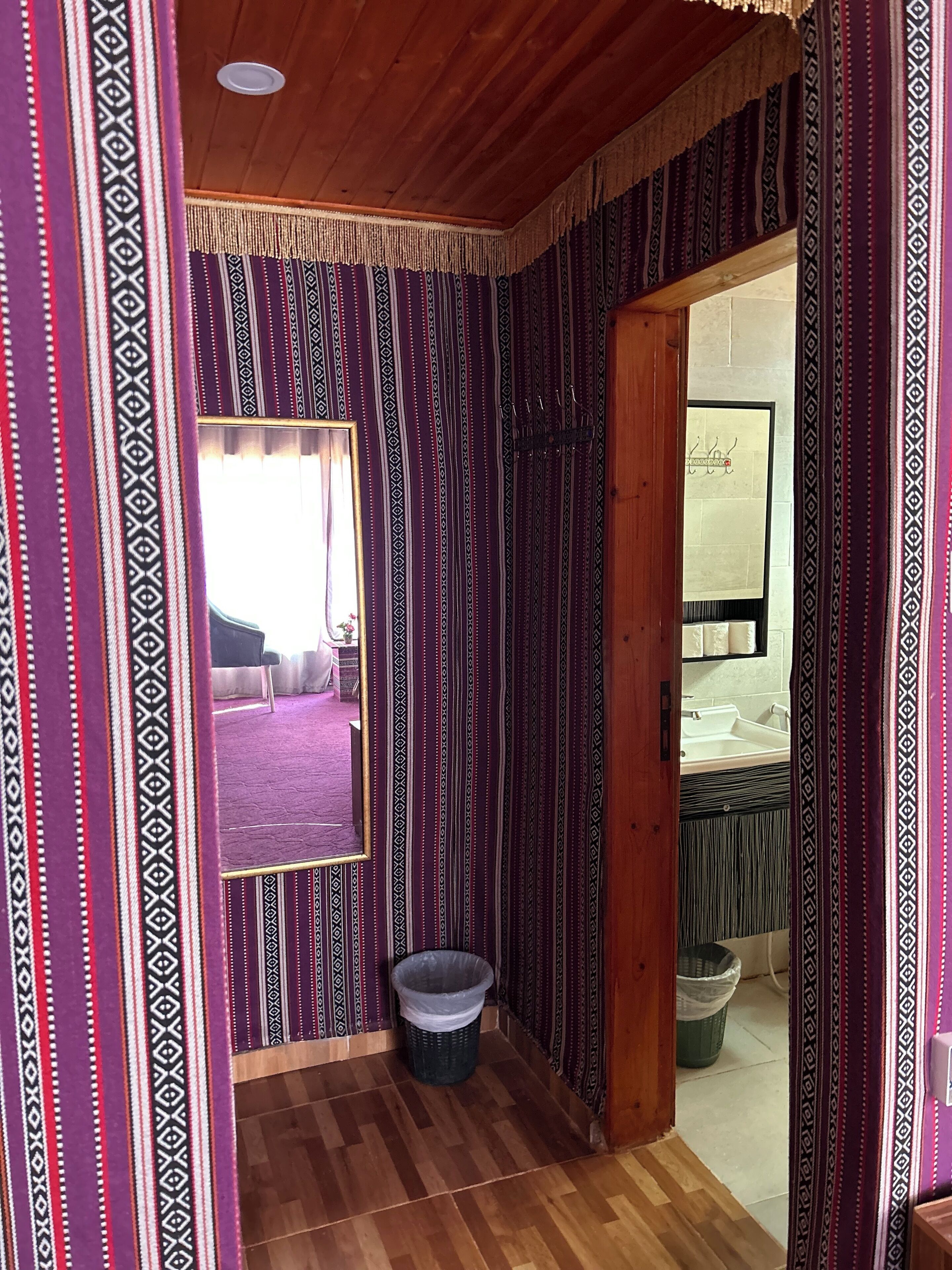 A room with patterned curtains, a wooden floor, a mirror, and a view into another room with a sink.