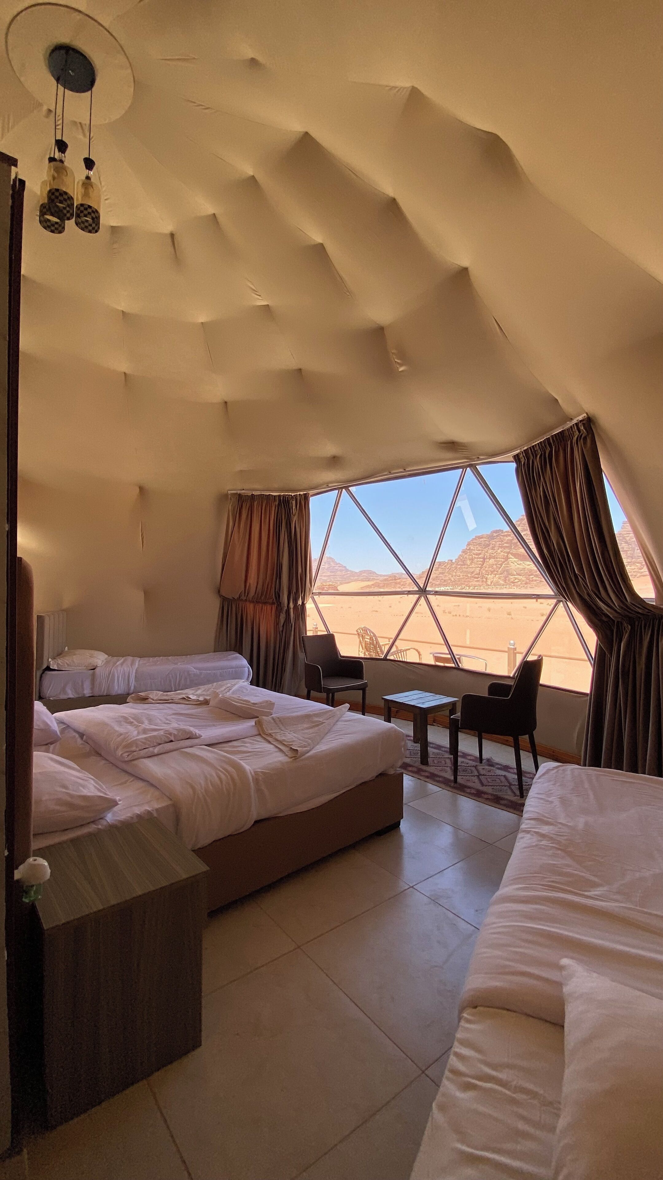 A spacious tented room with two beds, a small table, and a chair, featuring a large window with a desert view.