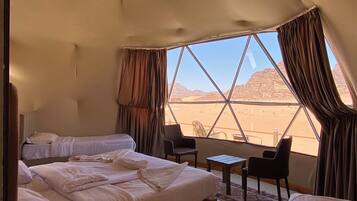 A spacious tented room with two beds, a small table, and a chair, featuring a large window with a desert view.