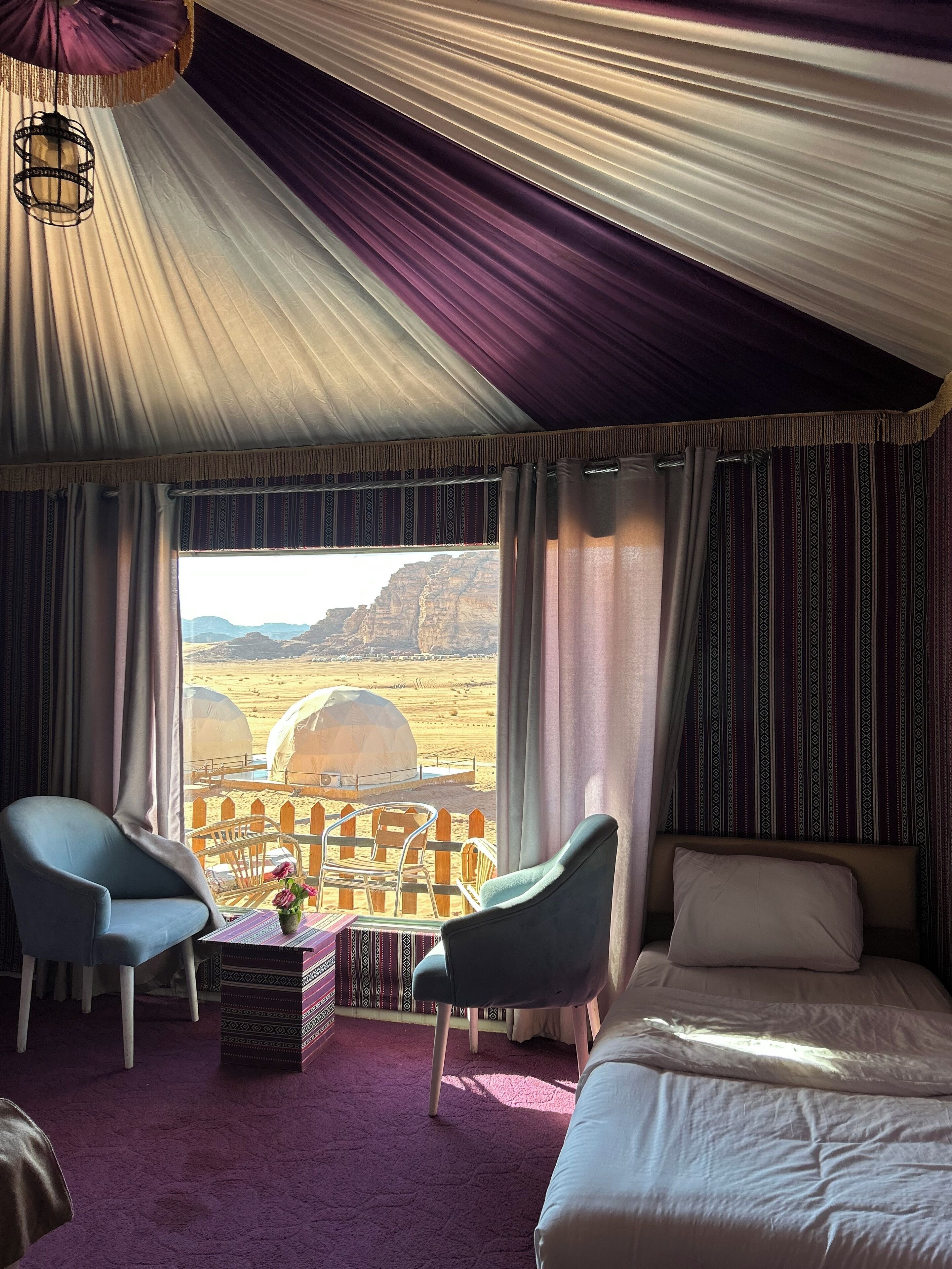 A room with a bed, two chairs, a small table with a vase, and a view of desert tents.