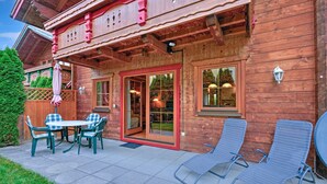 A wooden cabin with a patio featuring a table and chairs, a sliding glass door, and two lounge chairs. - Scenic Stay in Feriendorf Wildschonau Chalet (Wildschönau)