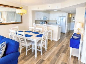 A modern kitchen with white cabinets, a central island, and a dining area with white chairs and blue placemats. - Ashworth 405 (North Myrtle Beach)