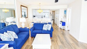 A modern living room with blue sofas, a white coffee table, and a kitchen in the background. - Ashworth #405! 4BR/3BA with Stunning Views & Prime Amenities (North Myrtle Beach)