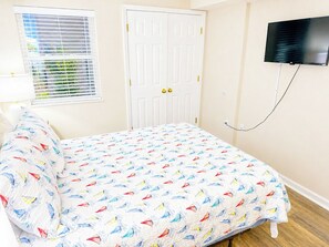 A bedroom with a bed, a white door, a window with blinds, and a television mounted on the wall. - Ashworth 405 (North Myrtle Beach)