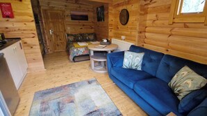 A wooden cabin interior with a sofa, a small table, a bed with a floral bedspread, and a kitchen area. - Snowdrop Lodge Dog Friendly Nr Anderby Creek (Alford)
