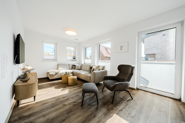 Un salon moderne avec un canapé, un fauteuil, un pouf et un téléviseur à écran plat fixé au mur.