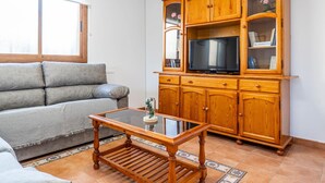 Un salon comprenant un canapé, une table basse avec un plateau en verre, un meuble en bois sur lequel est posée une télévision, et un sol carrelé.