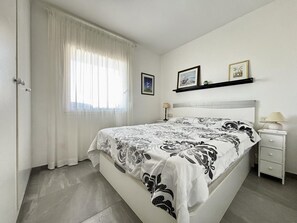 Un dormitorio con cama, mesita de noche, lámpara, ventana con cortinas y dos cuadros en la pared.