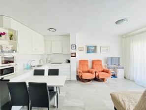 Une cuisine moderne avec des meubles blancs, une table à manger avec des chaises et deux fauteuils orange dans l’espace salon.