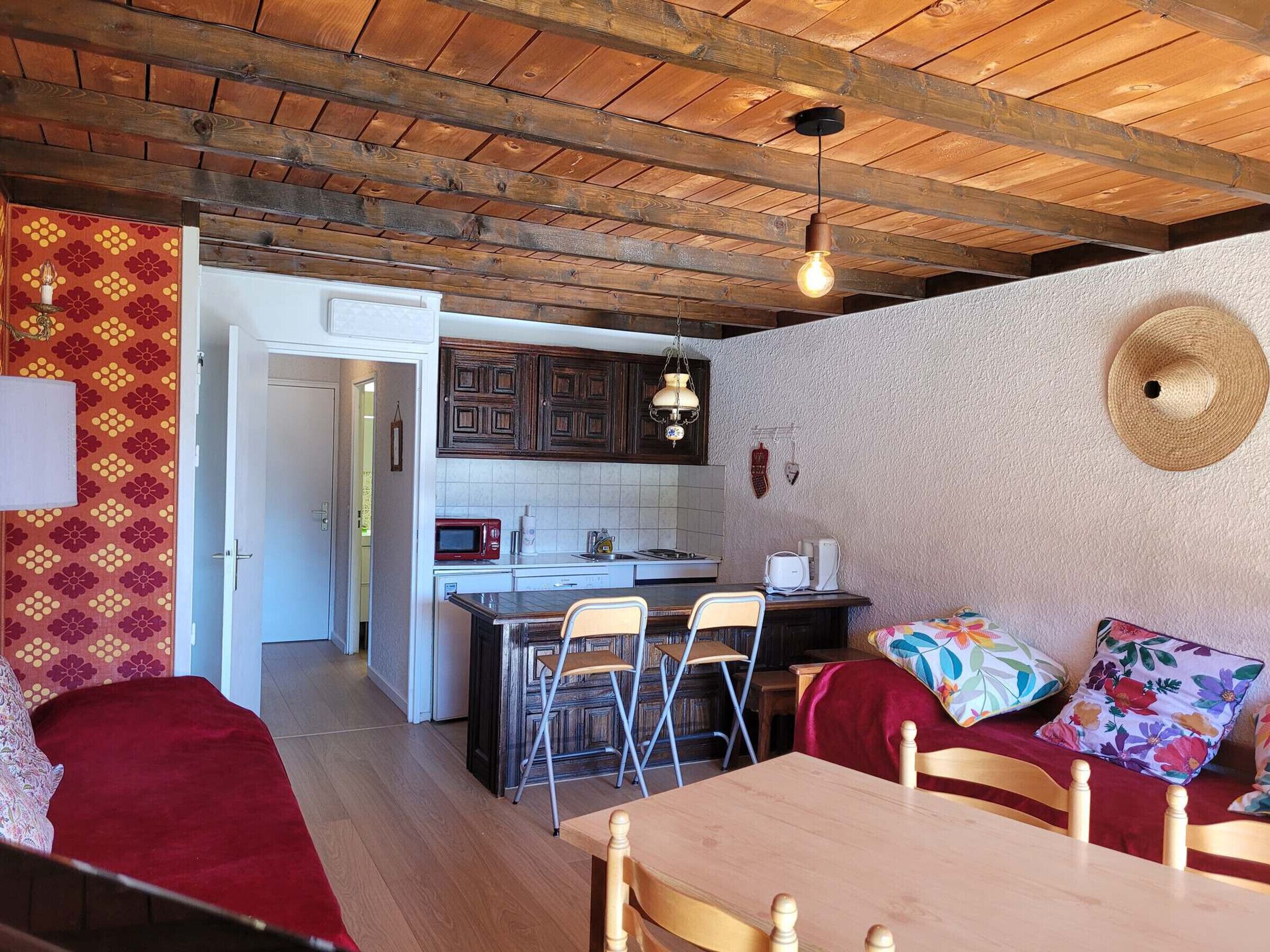 A cozy interior with a wooden ceiling, a kitchen area with a microwave and stove, a dining table with chairs, and a patterned wall.
