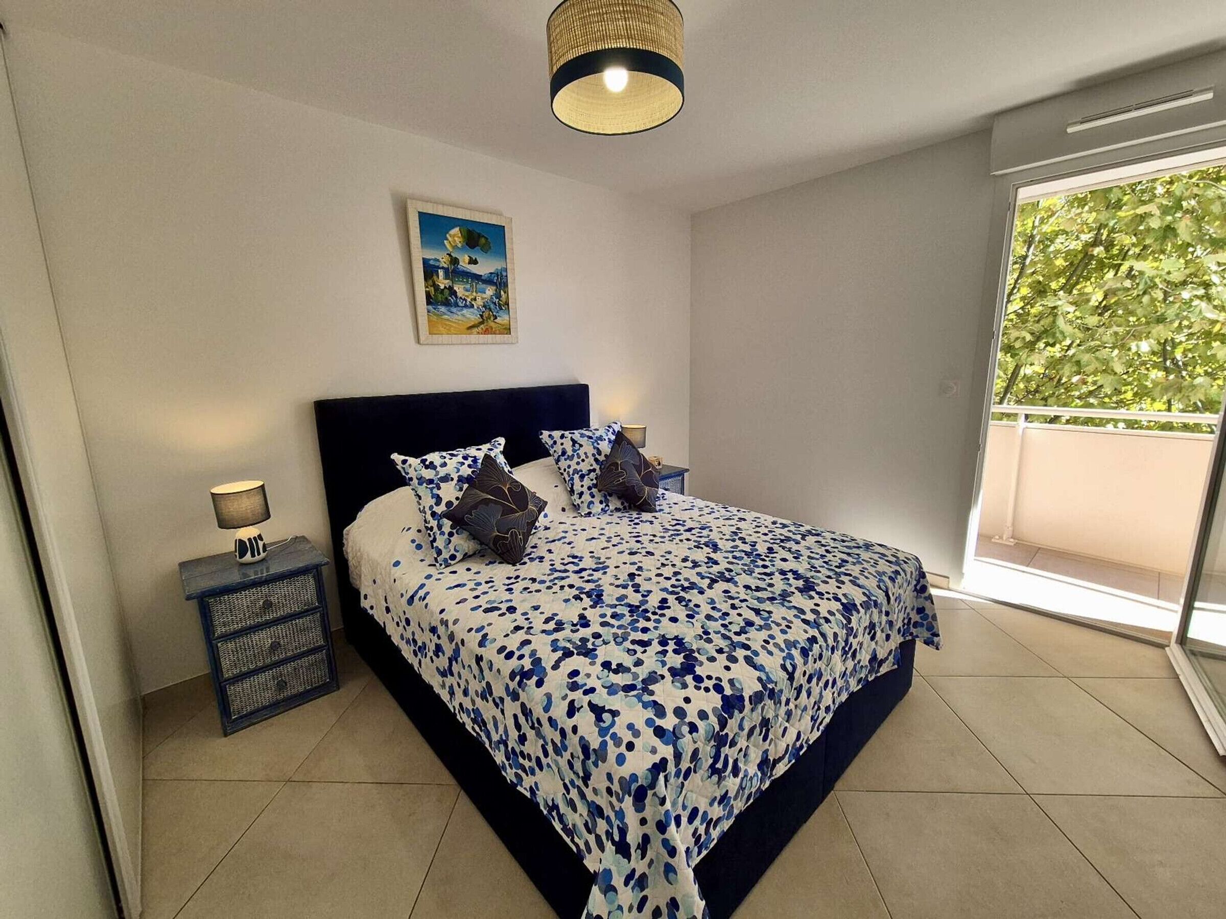 Un dormitorio con cama, mesita de noche, lámpara, un cuadro y vista a zonas verdes por la ventana.