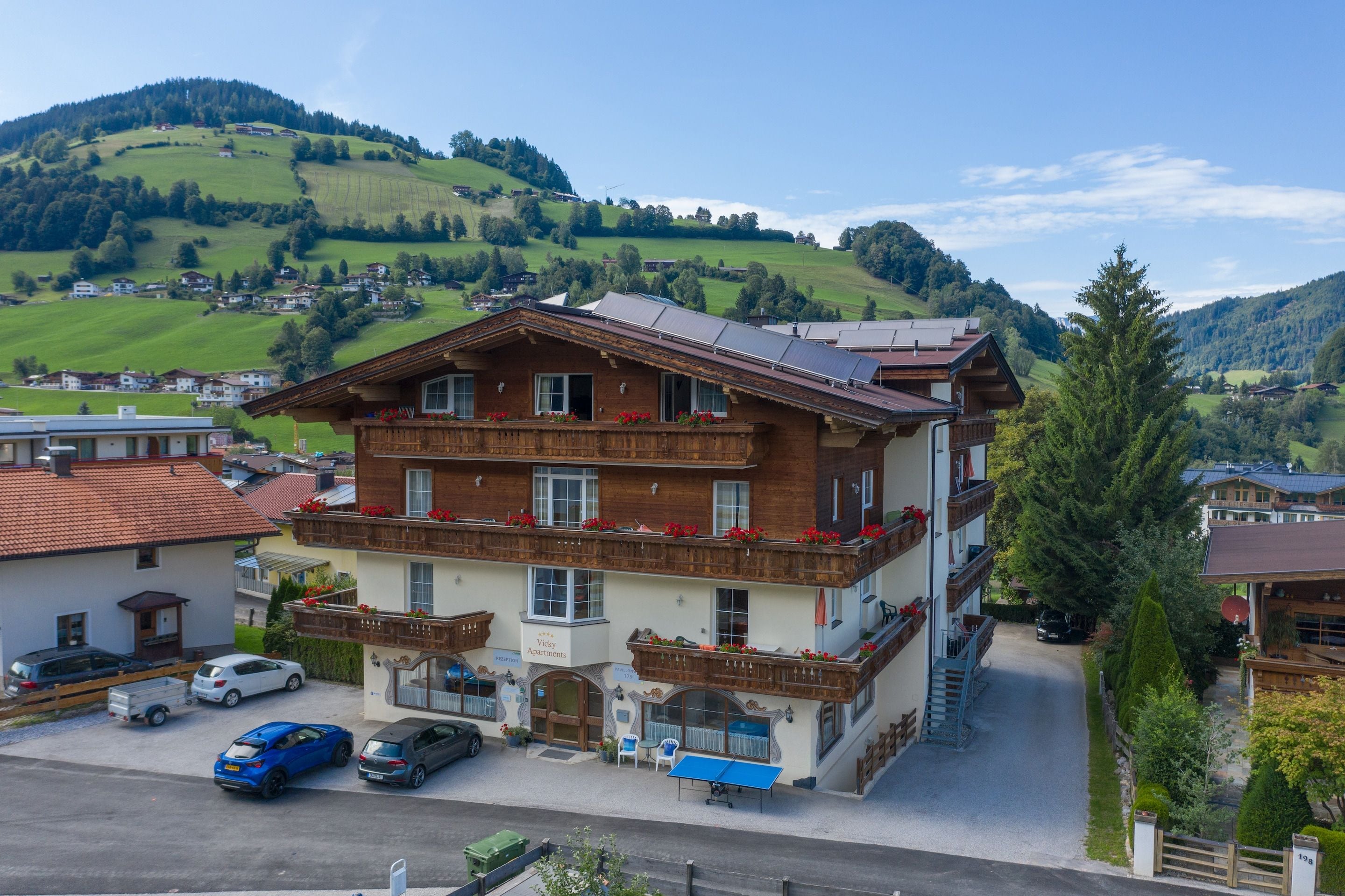 Un immeuble à plusieurs étages, de style architectural traditionnel, doté de balcons ornés de jardinières fleuries, d’un hall d’accueil et d’un parking où sont garées des voitures.