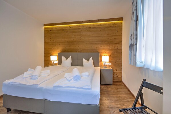 A double bed with white linens and towels, wooden headboard, bedside lamps, and a bench by the window. - Mountain View Ski Brunner Apartment Top 1 (Wildschönau)