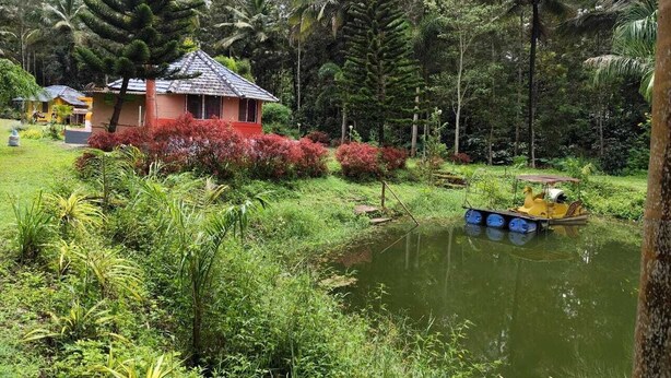 A house with a tiled roof, surrounded by lush greenery and a small pond with a boat. - Rising Surya Homestay (Kushalnagar)