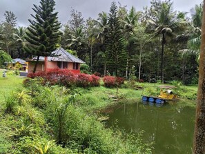 A house with a tiled roof, surrounded by lush greenery and a small pond with a boat. - Rising Surya Homestay (Kushalnagar)