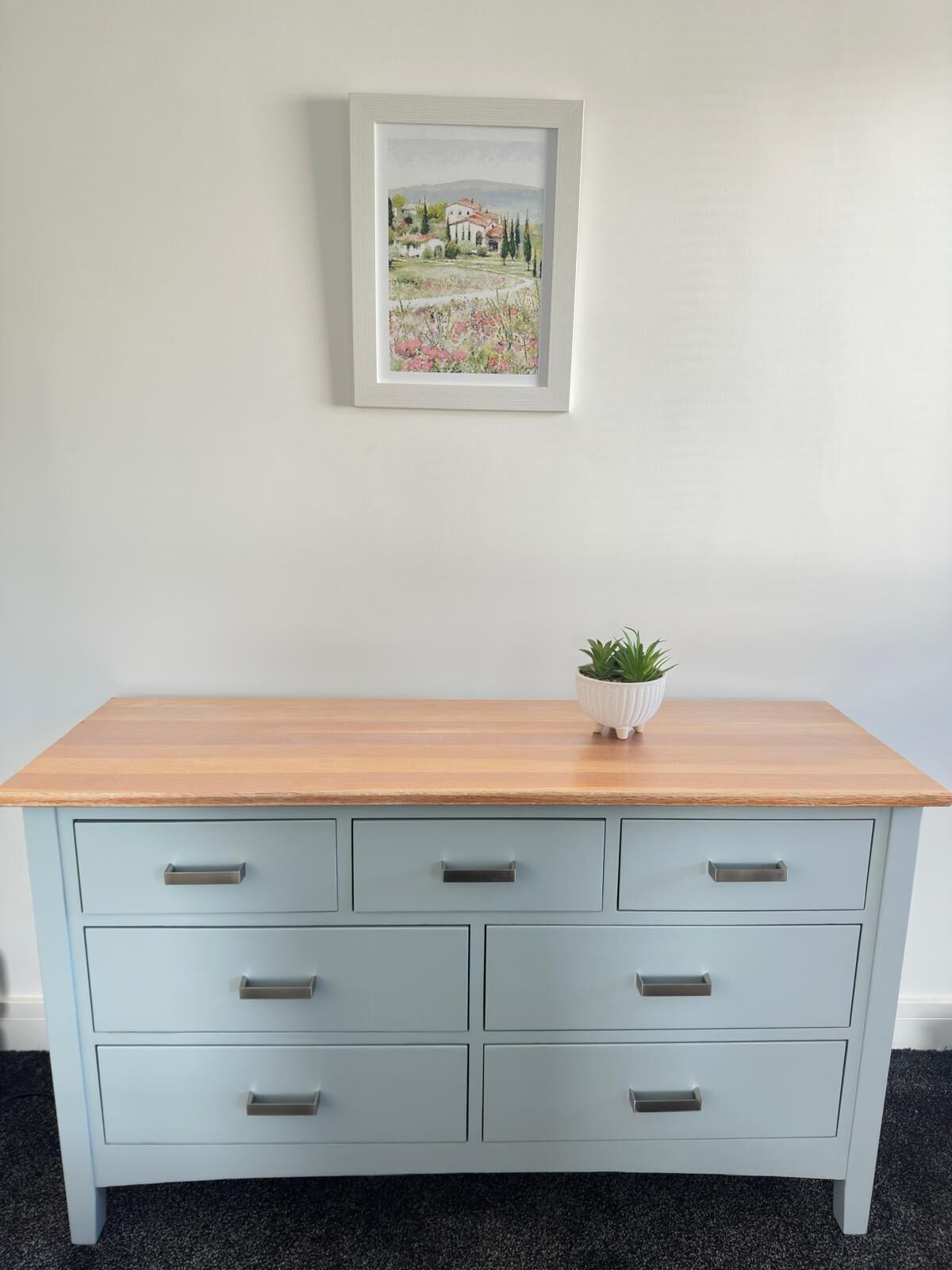 Een lichtblauwe dressoir met zes lades en een houten bovenblad. Daarboven hangt een lijst met een landschap.