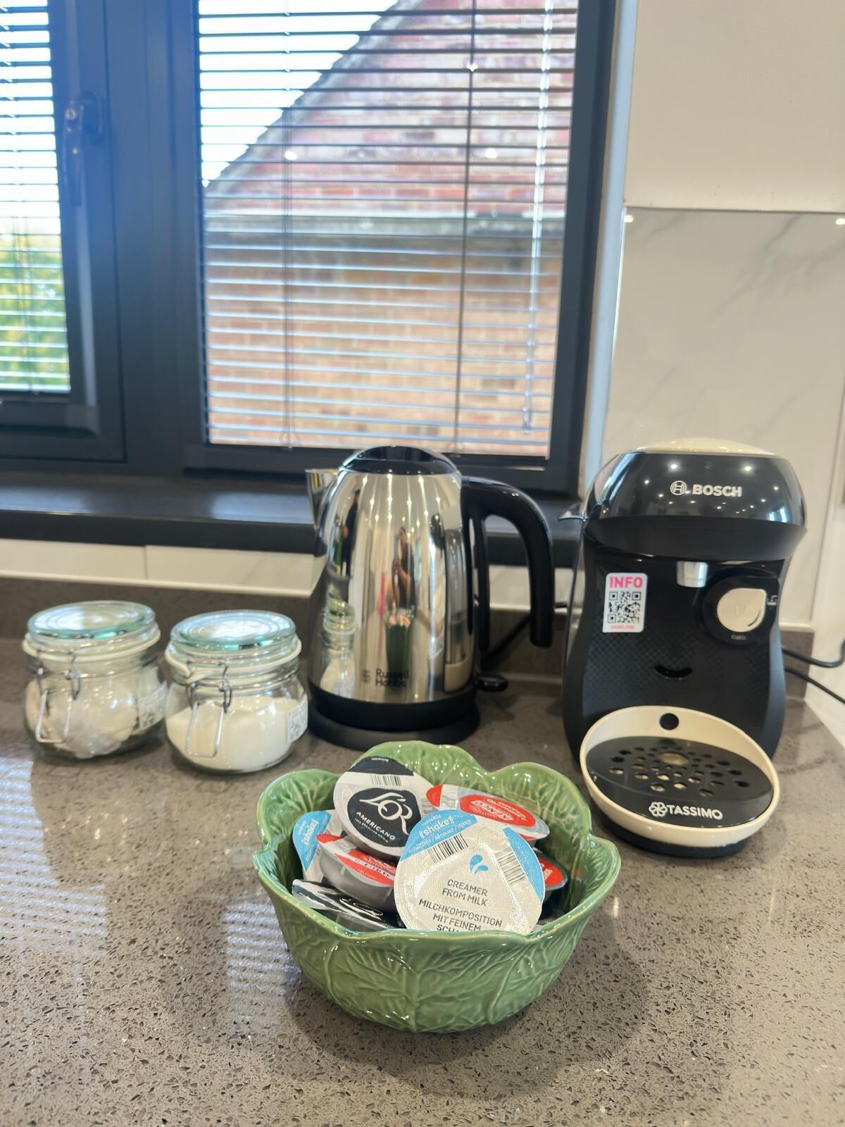 Un plan de travail de cuisine avec une machine à café, une bouilloire, deux pots de sucre et un bol de produits laitiers.