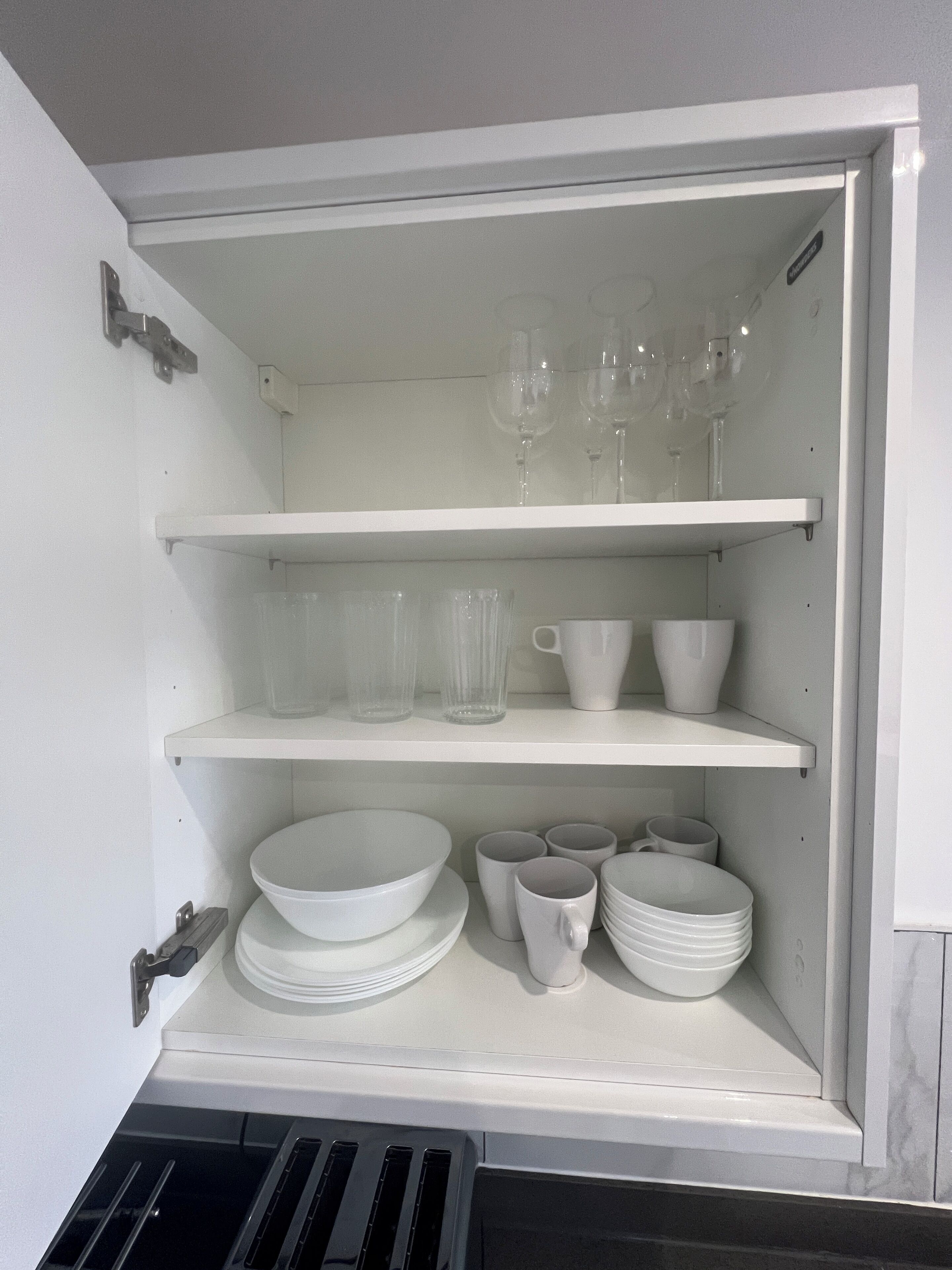 A white cabinet with shelves containing white dishes, glasses, and a white mug.