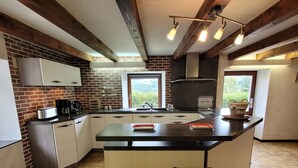 A modern kitchen with a large island, wooden beams, and a view of greenery. - Authentic Vosgian farm, sauna, and beautiful view (LA BRESSE)
