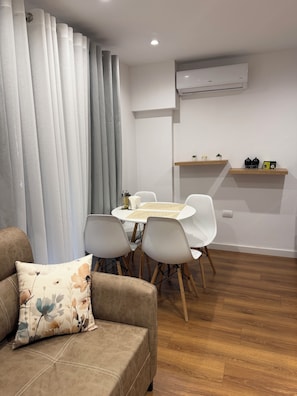 Ein modernes Wohnzimmer mit einer Couch, einem Esstisch mit Stühlen, einem grauen Vorhang und einer Wand-Klimaanlage.