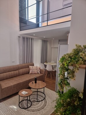 En moderne stue med en brun sofa, to små runde borde, et hvidt spisebord og en trappe, der fører til en etage ovenover.