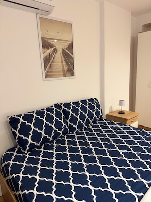 En seng med et geometrisk mønstret sengetæppe, et natbord med en lampe og et billede af en bro i ramme på væggen.