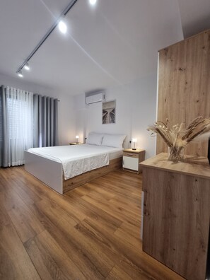 A modern bedroom with a wooden floor, a bed with white linens, a nightstand, a wooden side table, and a wall-mounted air conditioner.