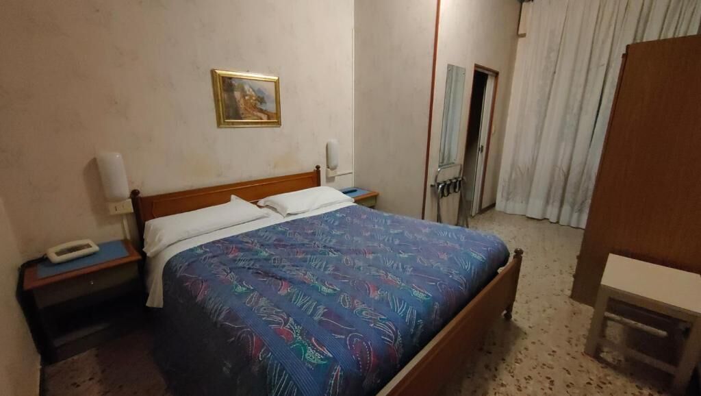 Et hotelværelse med en seng, et sengebord, et telefonbord, en telefon, et billede på væggen, et bruseområde og et gardin.