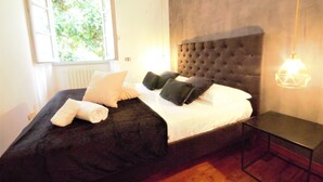 A bedroom with a large bed, a window with a view of greenery, a wall-mounted lamp, and a bedside table. - Villa Magnolia Como (Como)